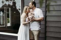 A family of three cuddle outside their home with their newborn baby.