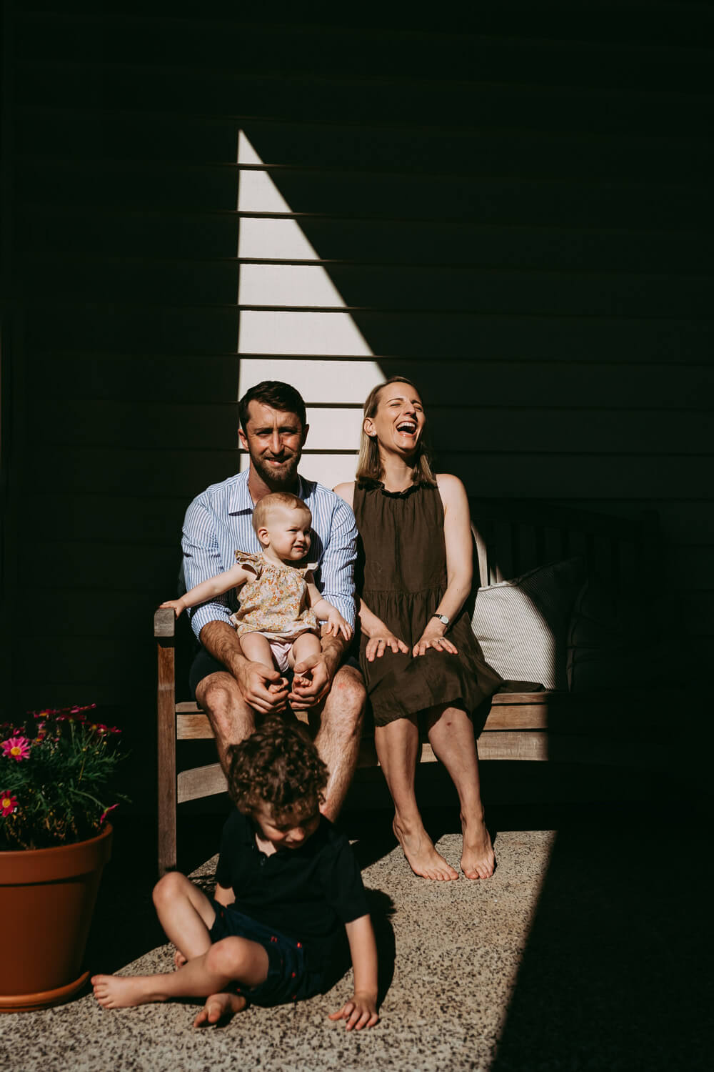 Murphy Family A family of four sits on a wooden bench. A woman is laughing as she throws her head back. They sit in a triangle of light, dark shadow surrounds them.