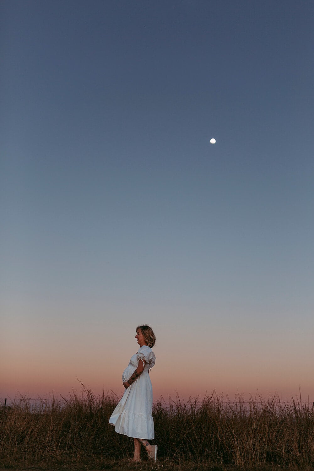 Murphy Family A pregnant woman wearing a white dress holds her belly. The moon shines above her.