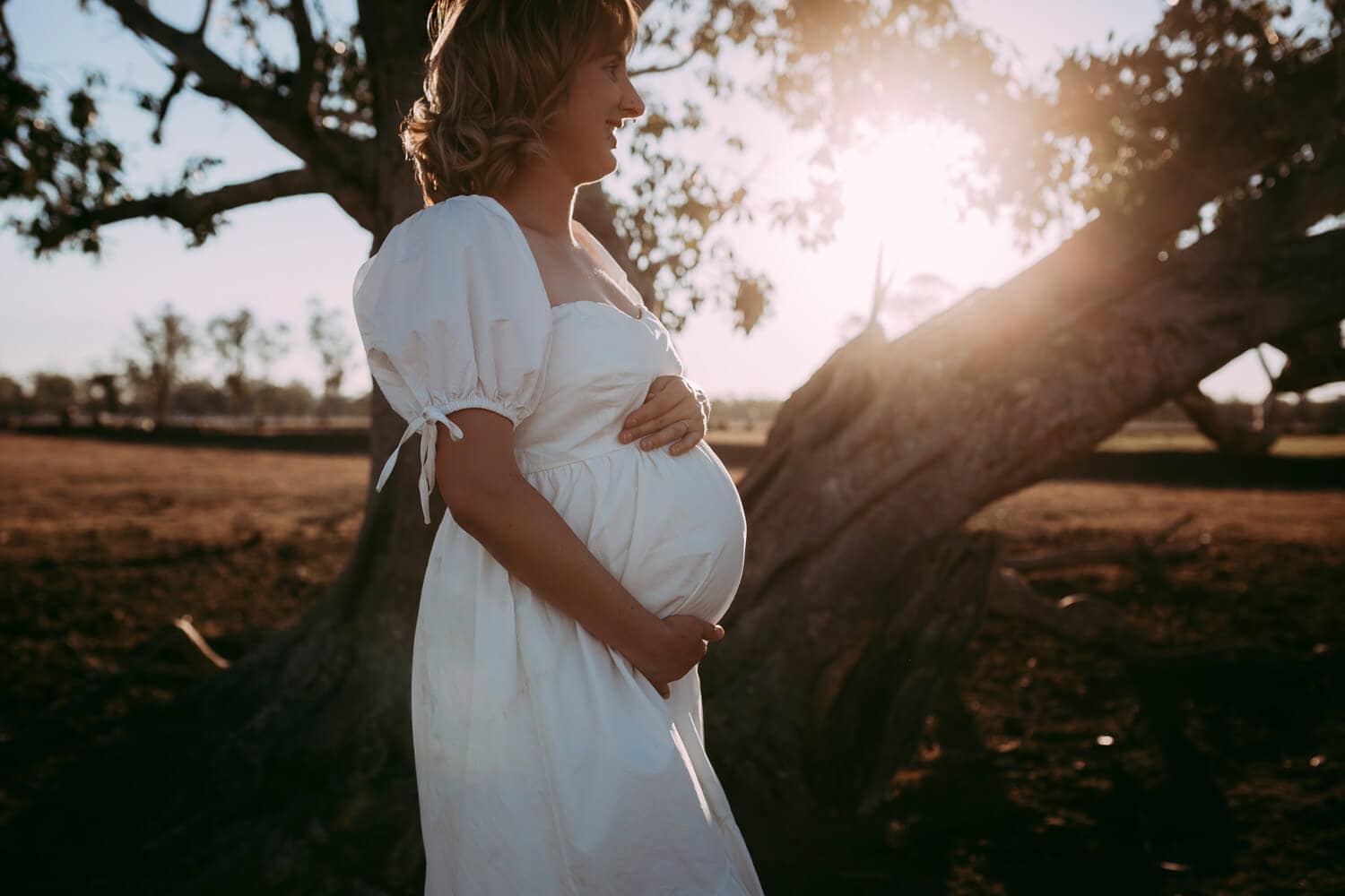Murphy family A pregnant woman wearing a white dress places her hand around her belly. A warm sun beams through the tree behind her.
