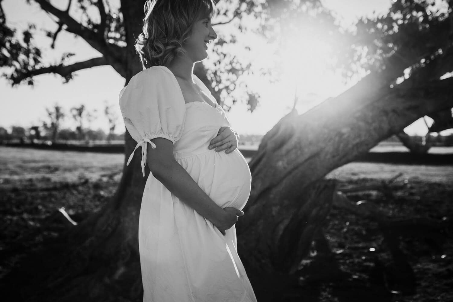 Murphy family A black and white image of a pregnant woman. She is wearing a white dress which contrasts against the dark background of a tree.