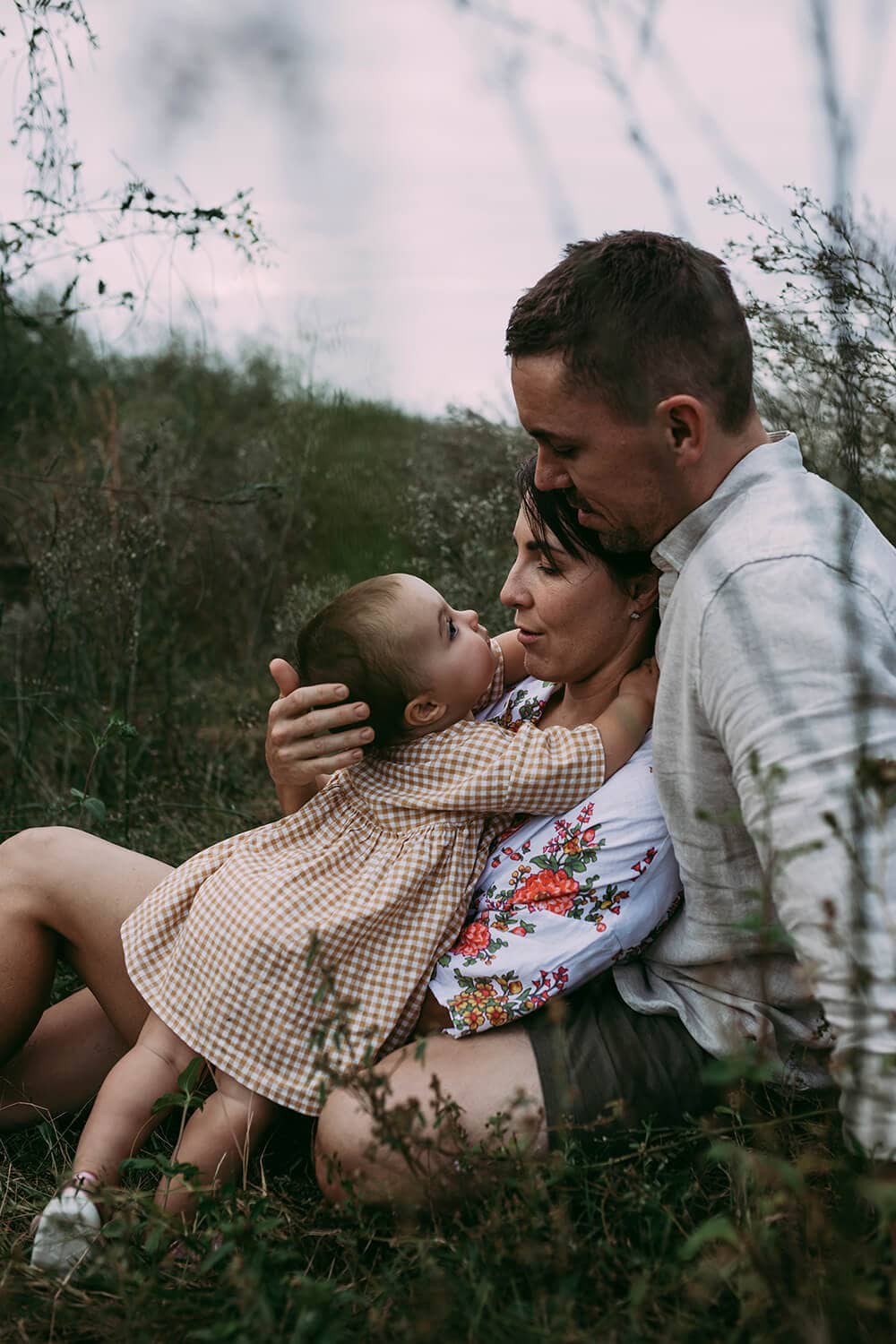 Murphy Family A toddler wraps her arms around her mother's neck as her father watches on. They're all sitting in a grassy field.