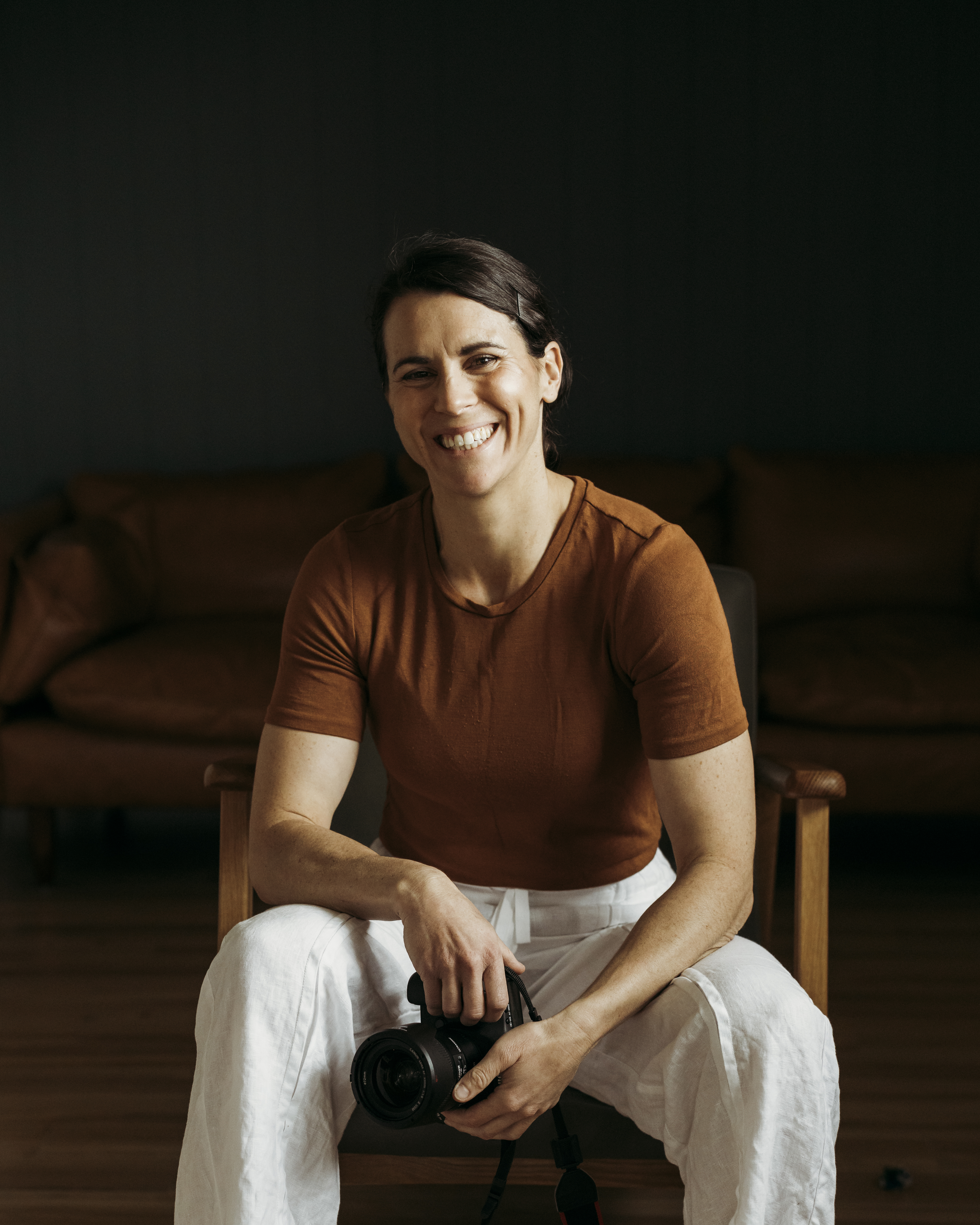 Alice self-portrait August 2023 001 crop A woman sits on a chair looking at the camera smiling. She wears a warm brown structured t-shirt and white linen pants. She casually rests both arms on her legs as she holds a camera.