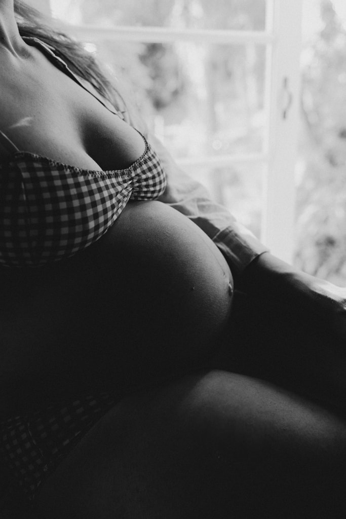 A black and white portrait of a woman's pregnant belly. Light from a window highlights the curve of her belly.