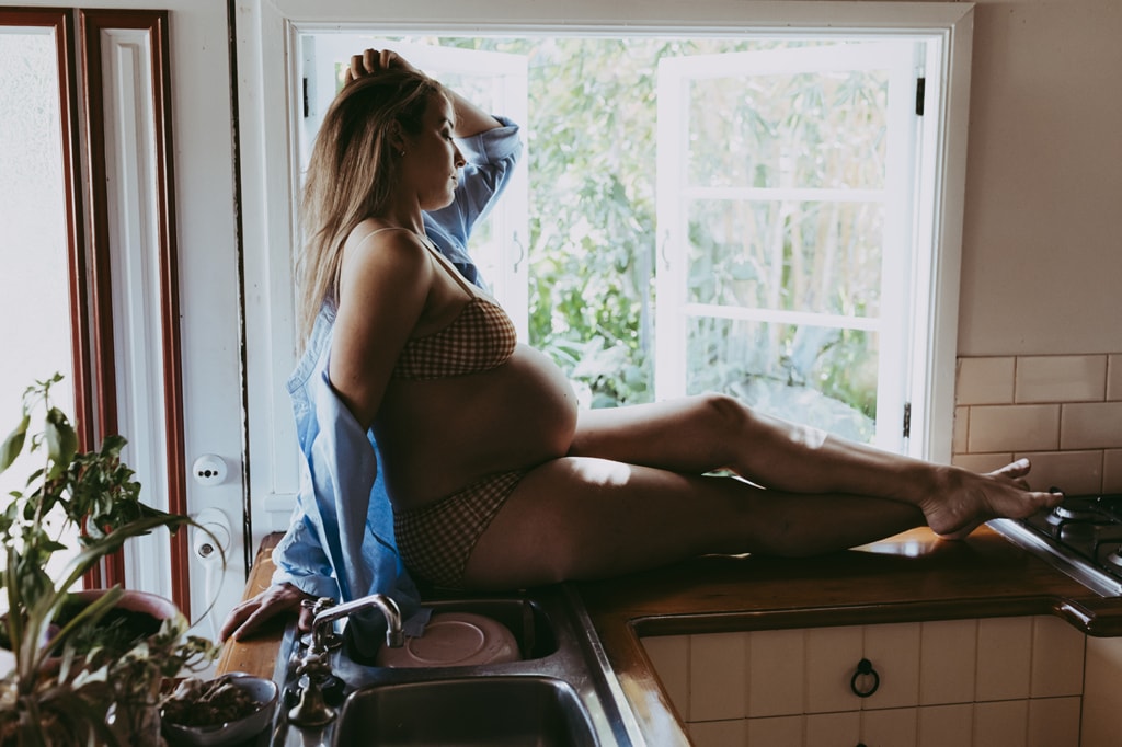 A woman sits on a windowsill. She's pregnant and is wearing undergarments with her belly showing. A blue shirt drapes across her arms.