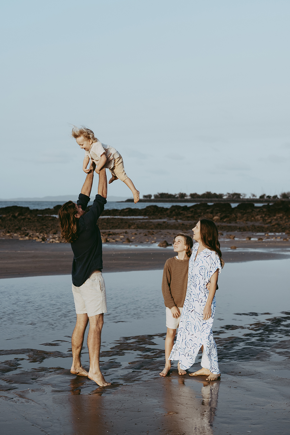 Family beach session 2 A family of four stands in shallow water on a beach. The father is throwing his young son in the air, as he giggles. While his mother and brother watch on as they stand together.
