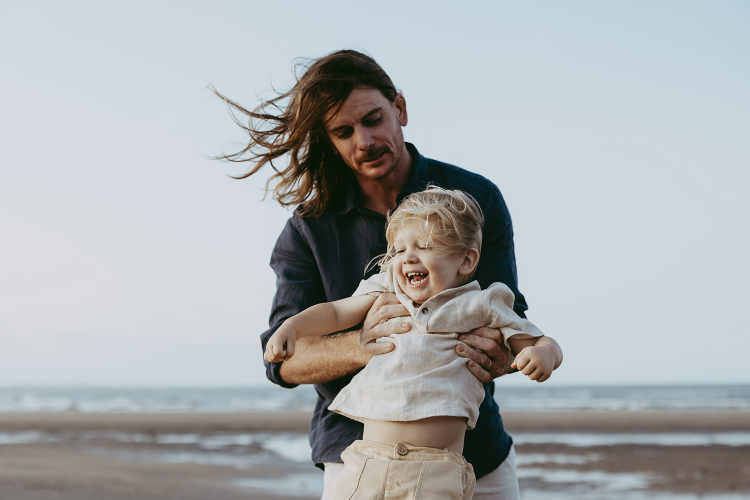 Lucy and Robbie Palmer October 2023 032_web A father and son play at the beach. The father holds his son in both hands as his son squeals with delight. Ocean and sand can be seen behind them.