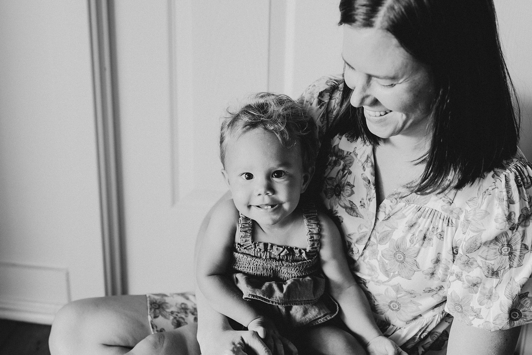 Naomi and Eden Buckley Jan 2024 003bw web A black and white image of a smiling toddler sitting on her mother's lap. Her mother is looking down at her smiling.