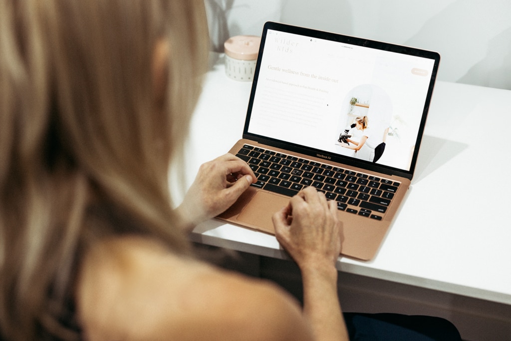 A laptop sits on a white desk with the Wilder Kids website on it. A woman sits at the laptop. You can only see her hands resting on the laptop and the back of her head.