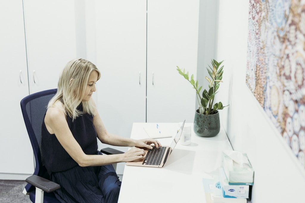 A woman sits at her laptop which sits upon a white desk.