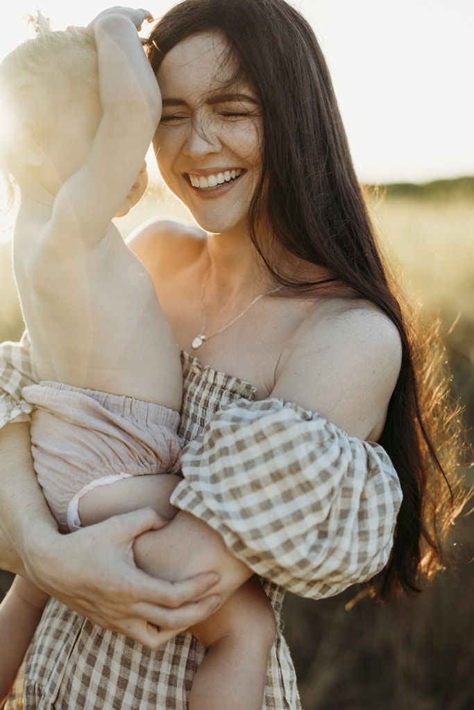 A mother laughs as she holds onto her wriggling toddler. The sun shines from behind them, illuminating her hair.