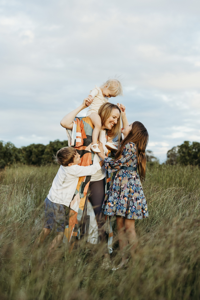 A mother holds her toddler on her shoulders while her two older children cuddle her. They are standing in a grassy paddock.