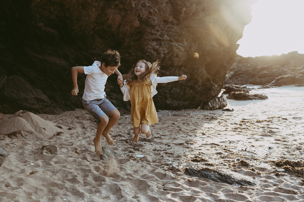 A boy and girl hold hands as they jump on a beach with a giant black rock behind them as the sun streams in.