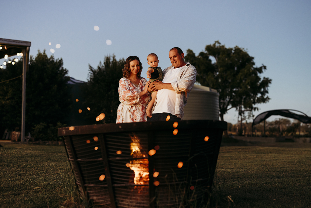 A family of three hold their toddler in their arms as they stand in front of a fire pit at their home. The sparks from the fire create little orbs of light across the photo.