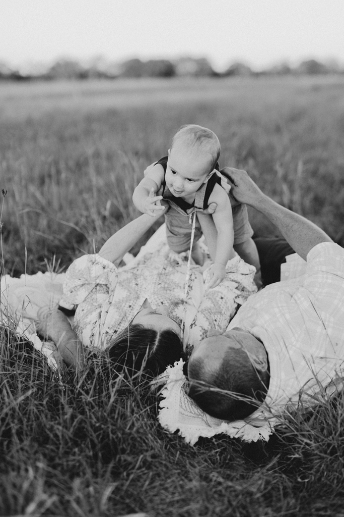 A toddler is held up by his parents who are lying on the ground in a grassy paddock.