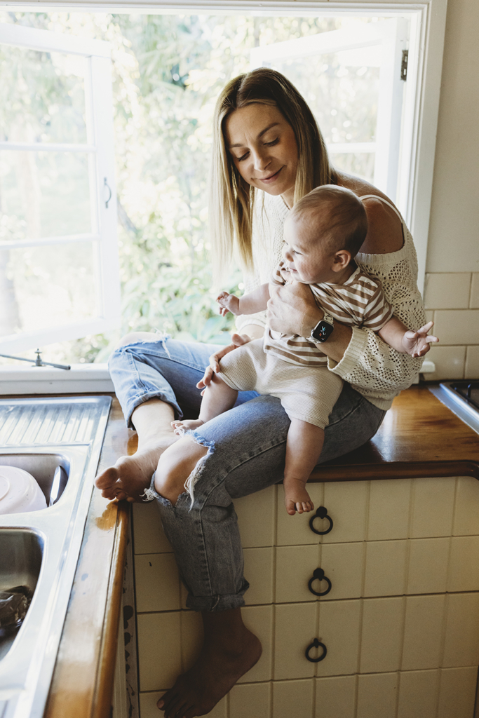 A mother holds her baby on one leg as she sits on a kitchen bench by a window.