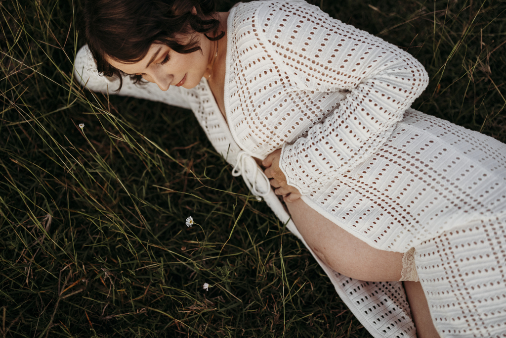 A pregnant woman lies in long green grass. There are three paper daisies near her. She is wearing a cream knit cardigan.