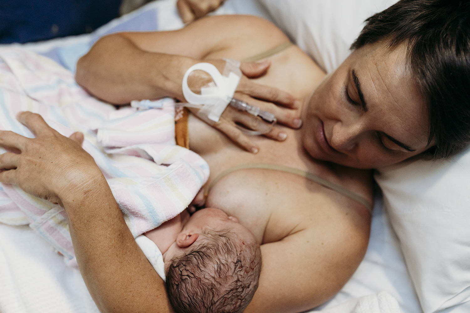 Mater Rockhampton newborn web A mother looks down at her newborn child while breastfeeding. The mother has a catheter in her wrist and is lying down.