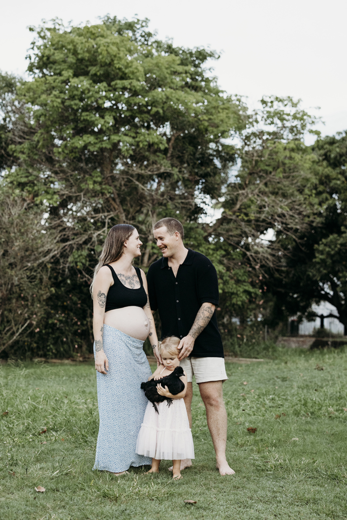A family of three stand in their backyard as they prepare to welcome a new baby. Their toddler holds their pet chook.