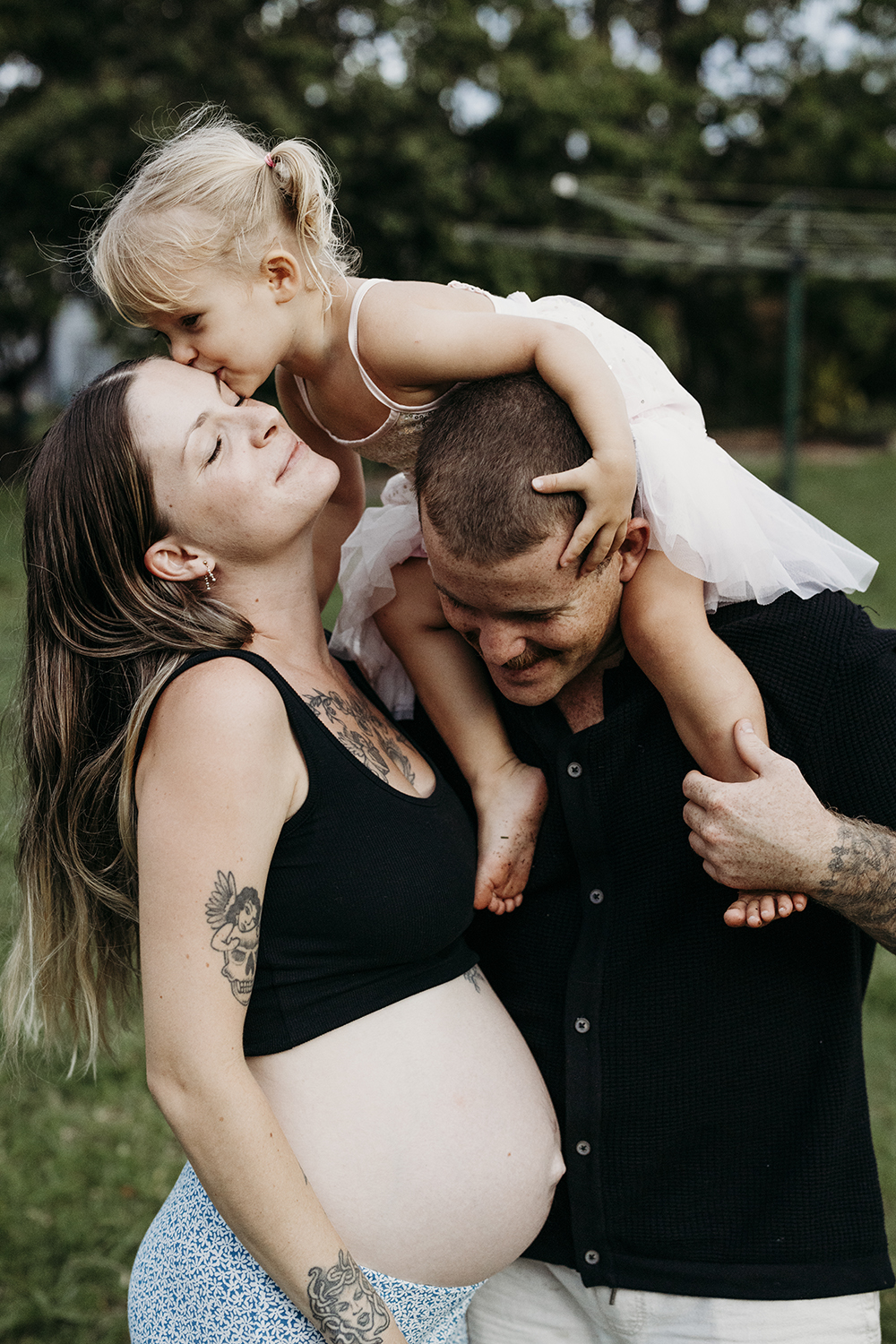 A family of three cuddle one another. A little girl sits on her father's shoulders and leans down and kisses her mother who's pregnant belly is showing.