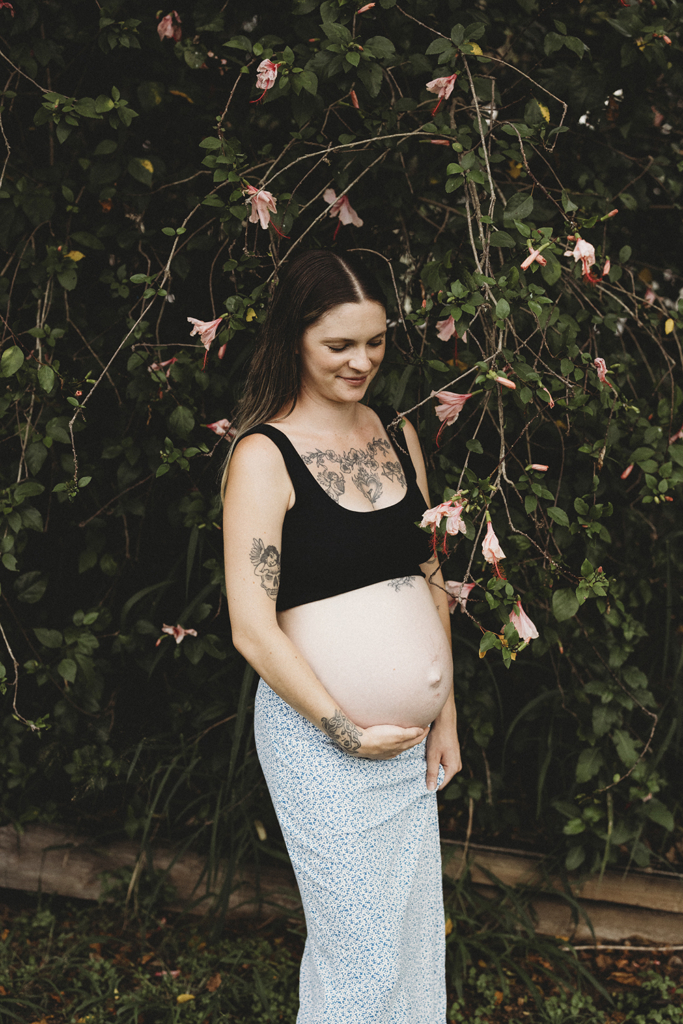 A woman stands smiling, gently placing a hand on her pregnant belly.