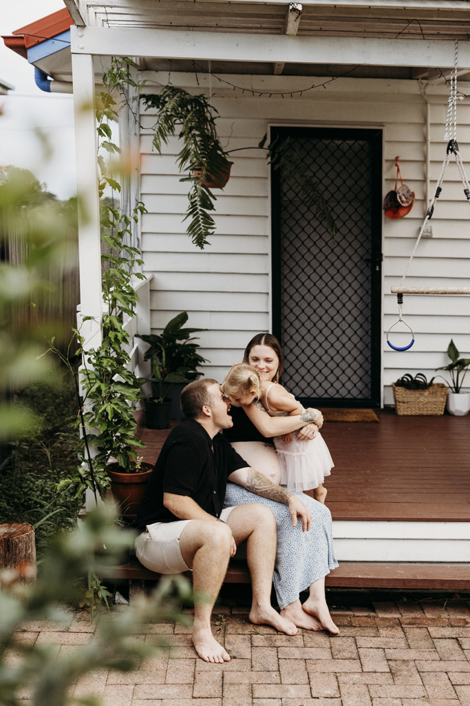 A family of three snuggle with one another on the front step of their home. The woman is pregnant.