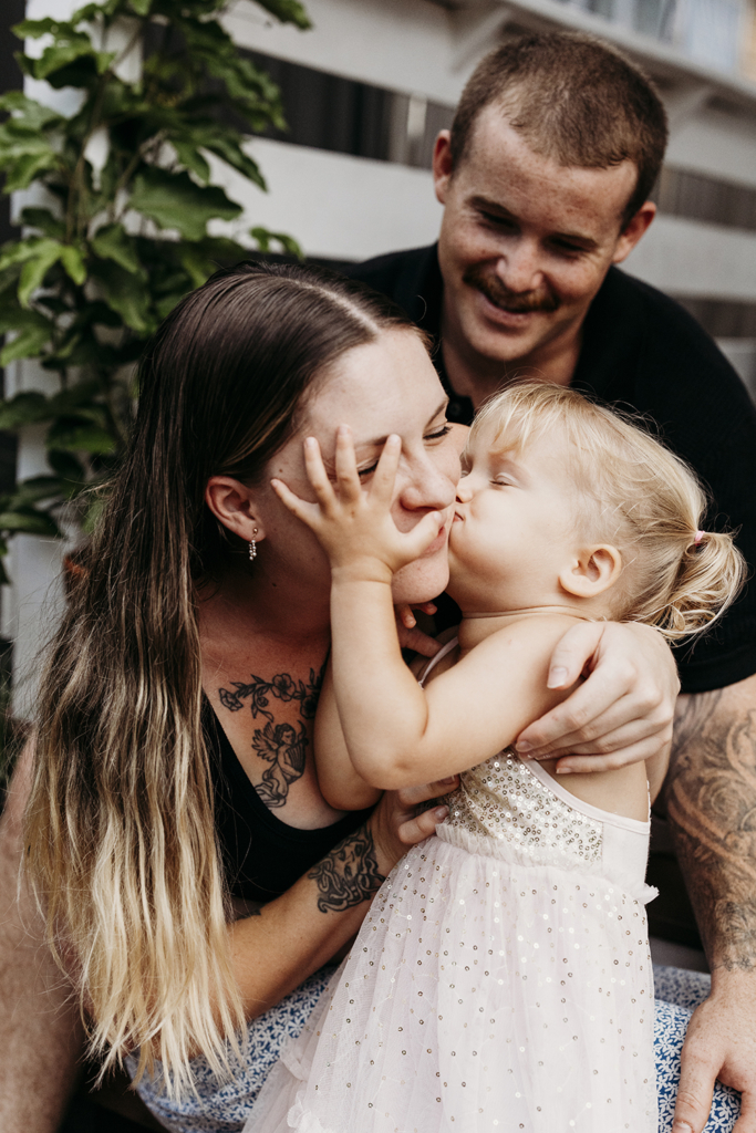 A family of three sit on their front steps as their toddler kisses their mother's cheek.