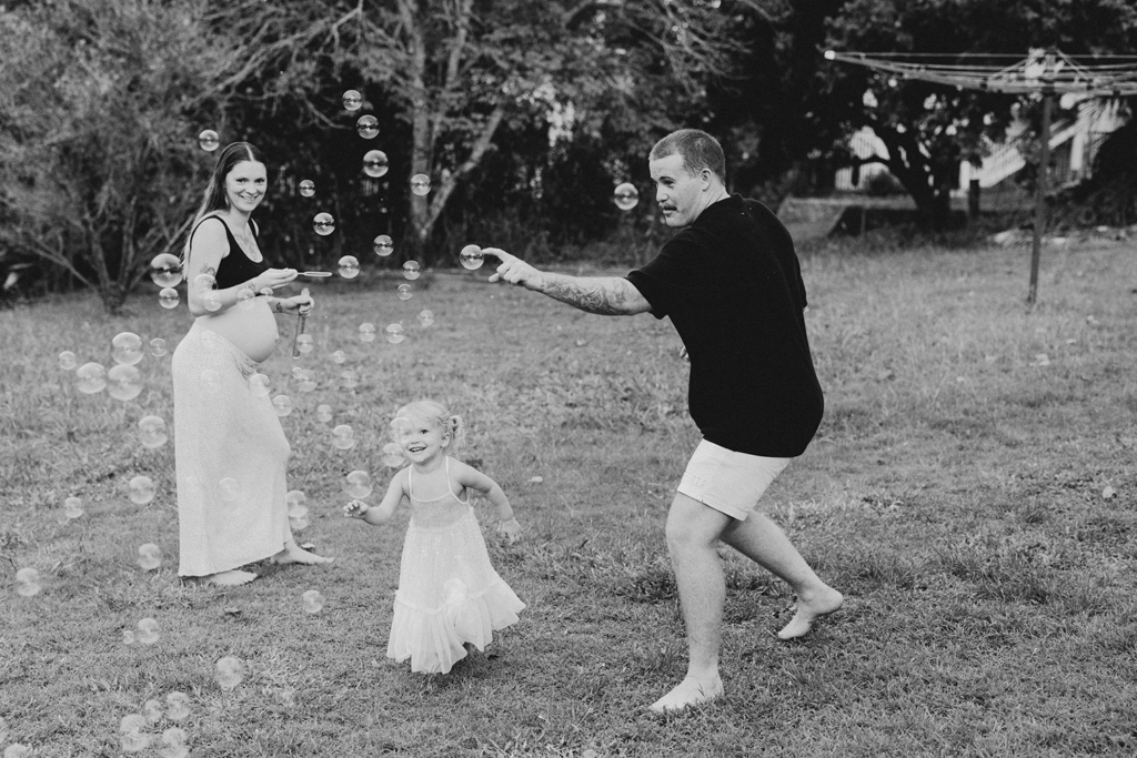A fun maternity session where a family of three blows bubbles in the backyard.