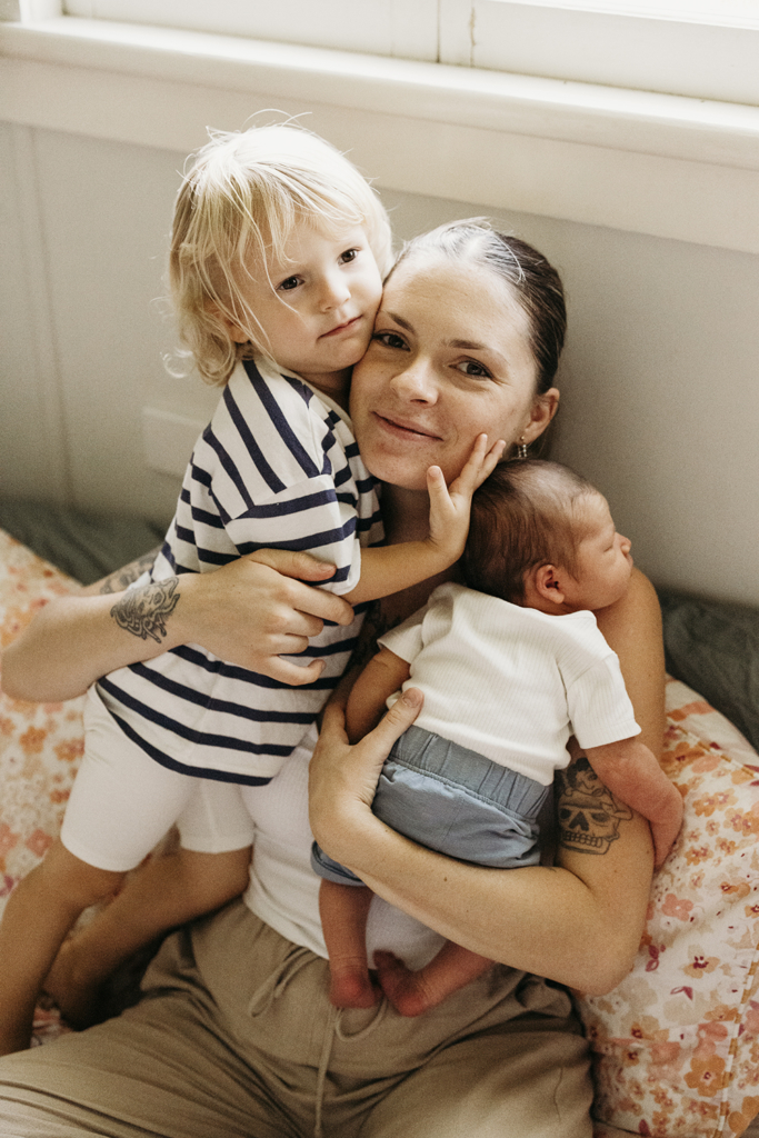 A mother cradles her newborn against her chest and cuddles her toddler.