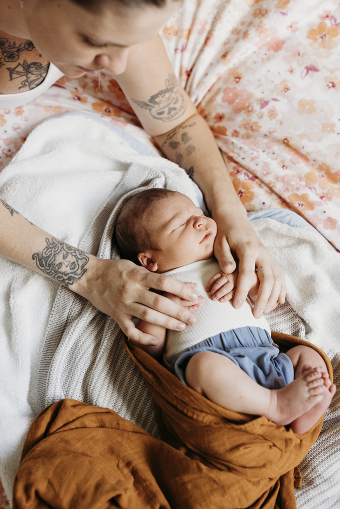 A mother holds the hands of newborn sleeping baby, as they lie in bed.