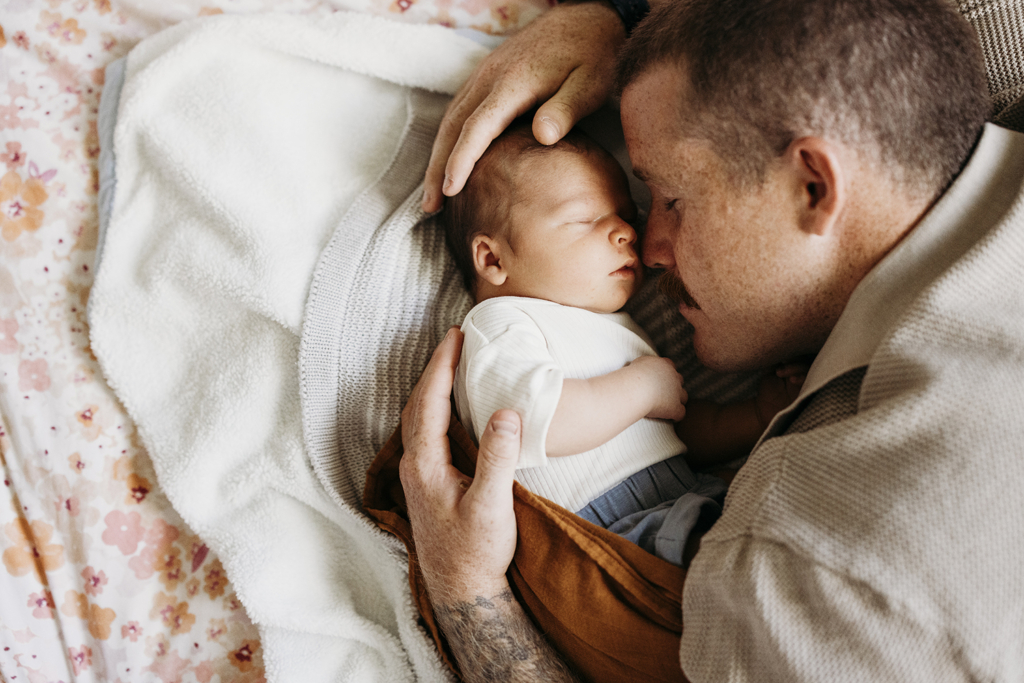 A father lies down next to his newborn son and snuggles him.