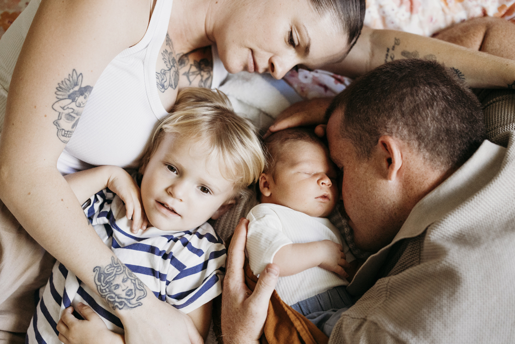 A family of four snuggle up on a bed. Mum and dad look down at their toddler and newborn baby.
