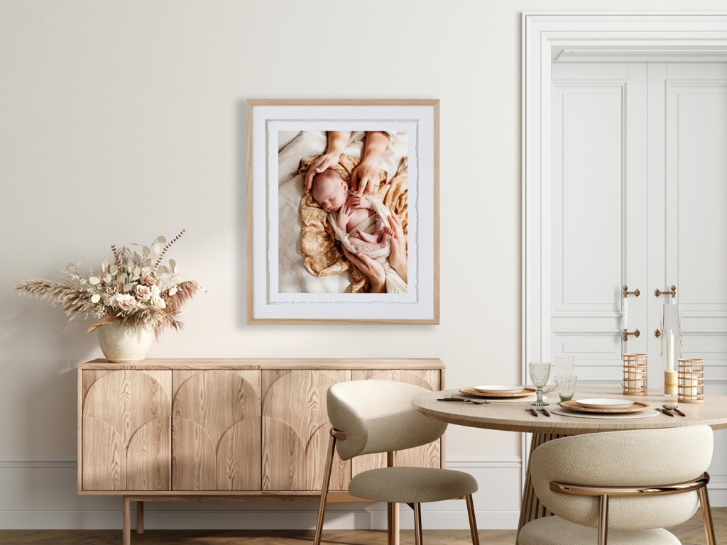 A fine art frame hangs in a dining room with a portrait of a newborn baby.