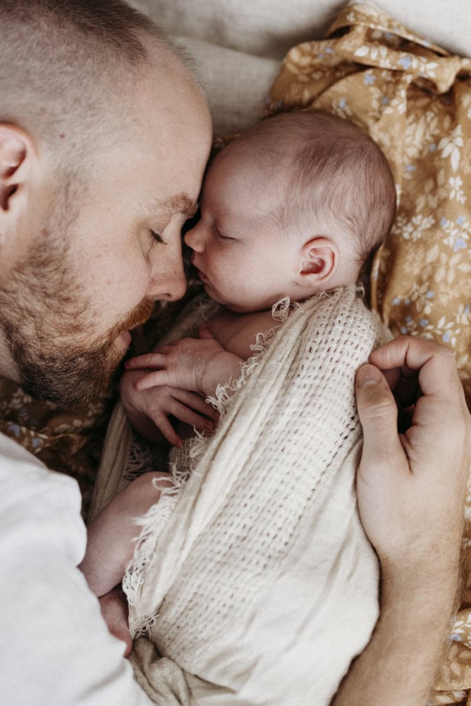 A father lies next to his sleeping newborn daughter. Both have their eyes closed.