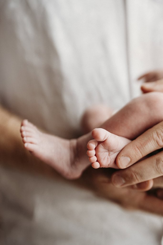 A father holds his newborn baby in a photography studio. His hands gently cradle her feet.