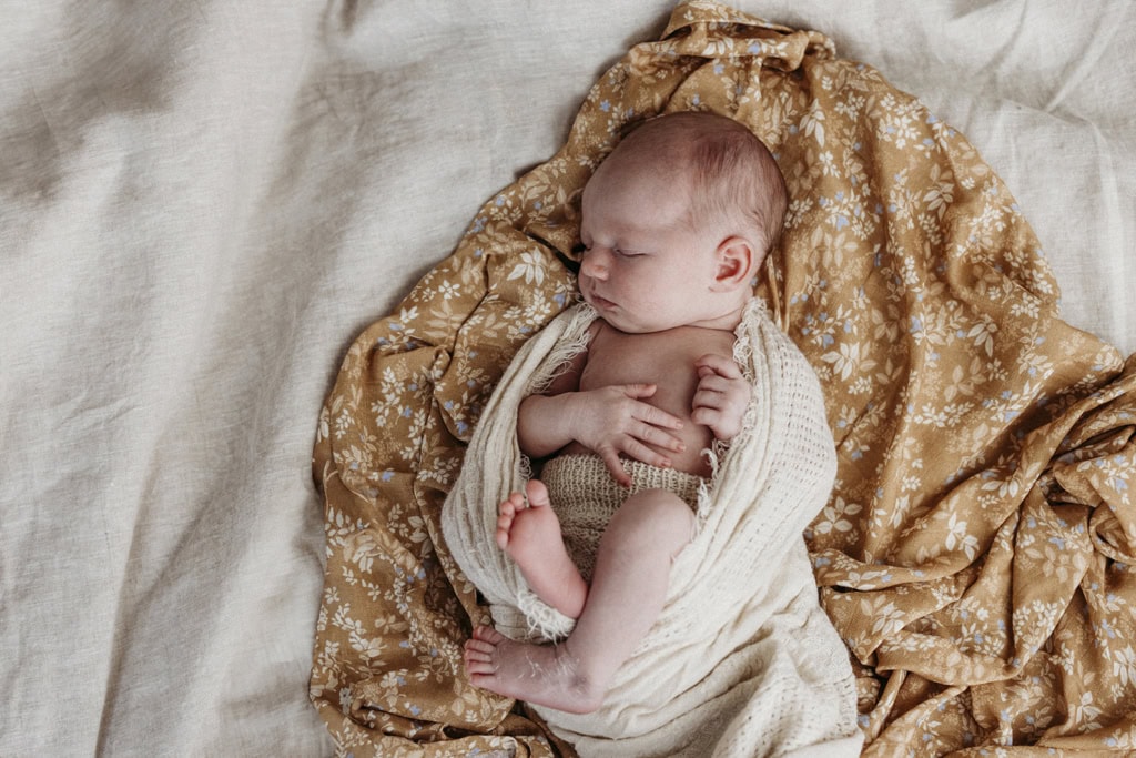 A newborn baby lays asleep. She's gently wrapped in a muslin cloth with her hands gently resting on her chest.