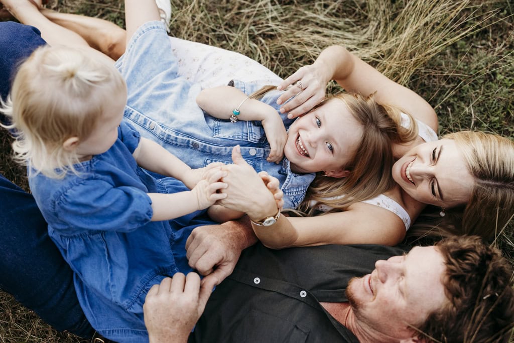 A young girl looks up at the camera smiling as she lies in long grass with her mother, father and toddler sister.