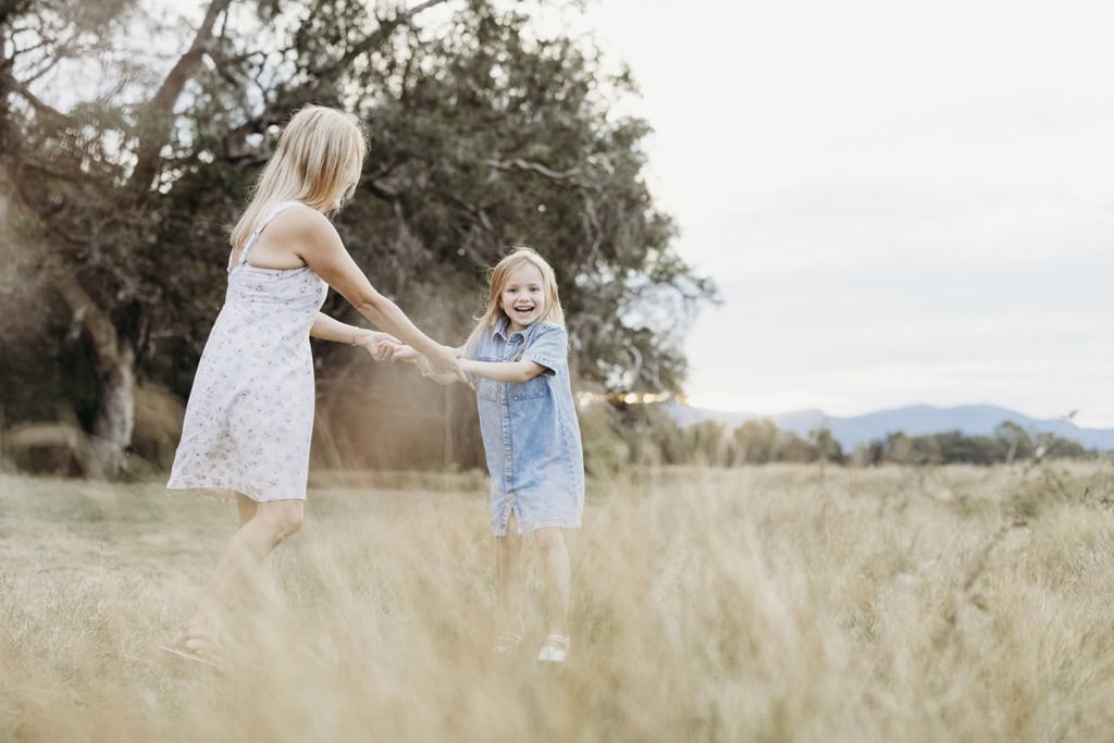 A little girl laughs as she twirls around with her mother in a paddock of long grass.