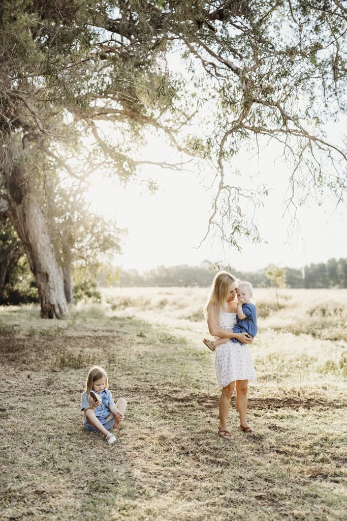 A mother stands as the sunlight streams through the trees behind her. She cradles her toddler as her other daughter, sits on the ground.