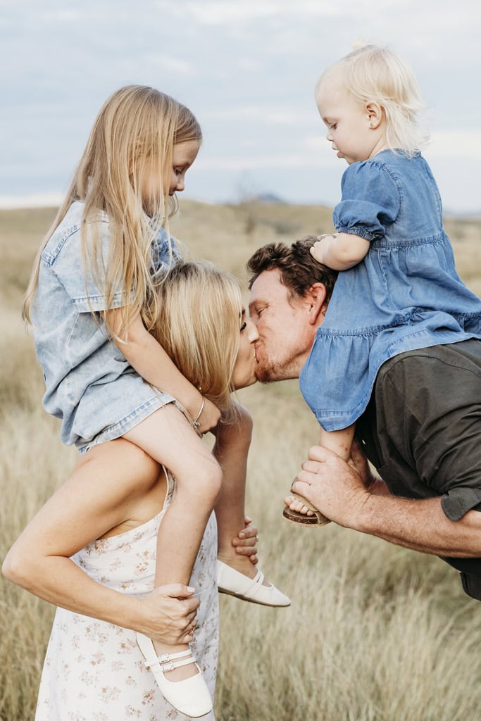 A couple kiss as their daughters watch on from their shoulders.