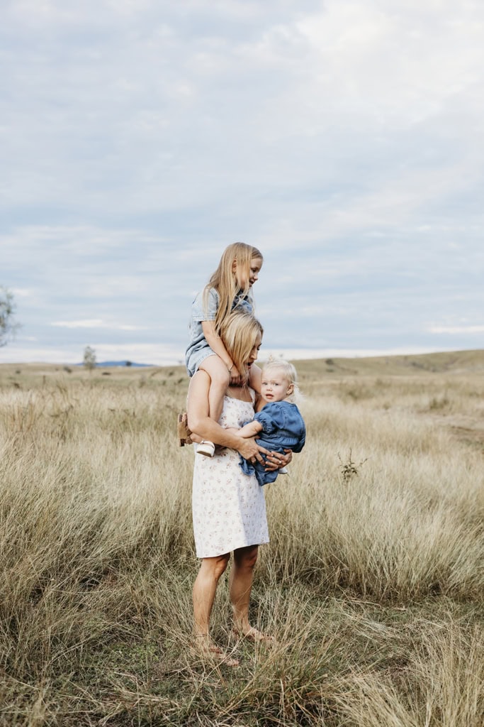 A mother holds one daughter on her shoulders as they carries her toddler in front.