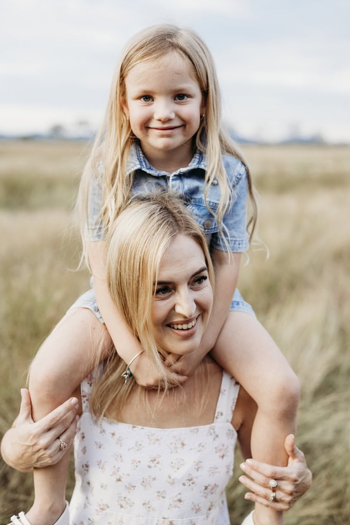 A mother smiles as she carries her daughter on her shoulders.