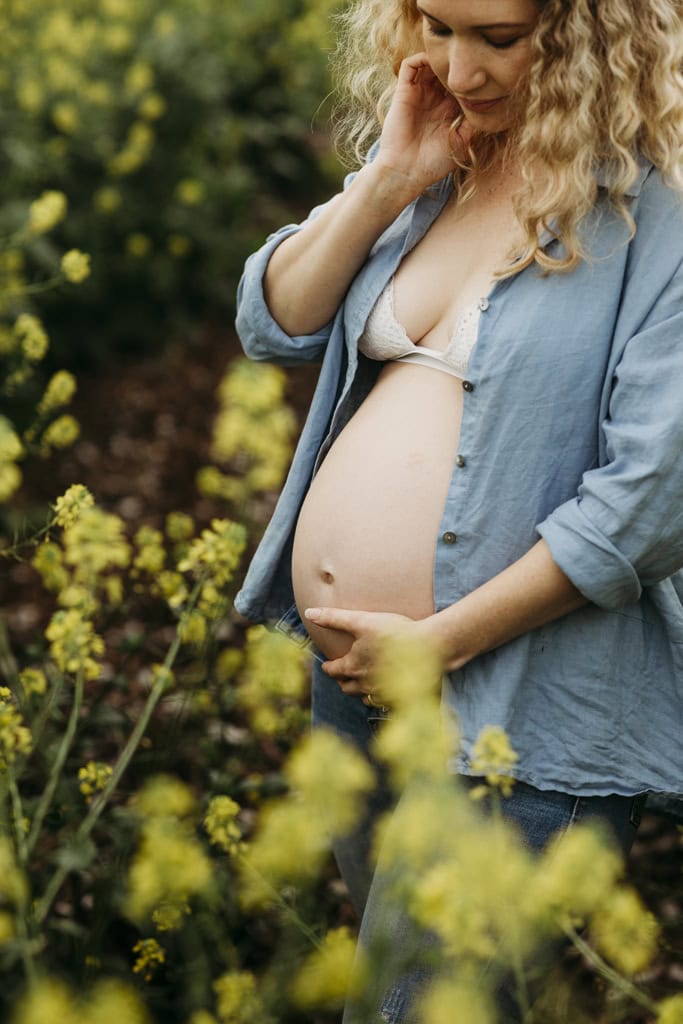 A woman looks down towards her pregnant belly as she stands in a field of yellow flowers.