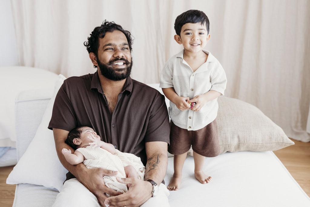 A father smiles as he holds his newborn and sits with his toddler son.