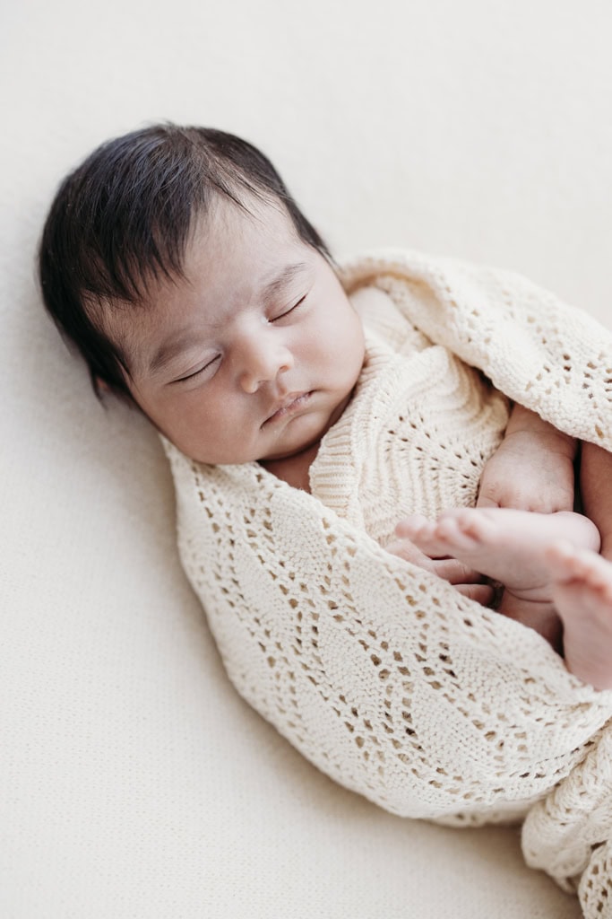 A newborn is wrapped in a neutral coloured blanket with her legs curled as she sleeps.