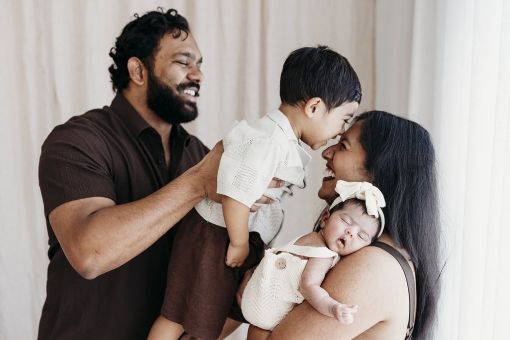 A family of four laugh as a mother cuddles her newborn daughter, and her partner holds their toddler son as he kisses his mother's head in a Rockhampton newborn photography studio.
