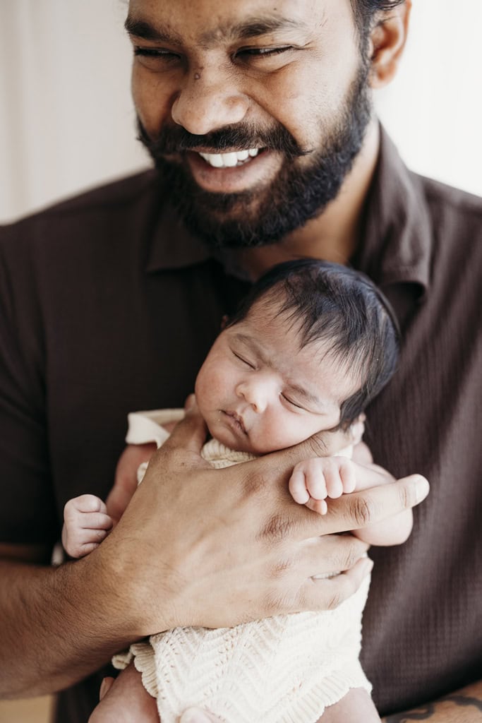 A father smiles as he holds his sleeping newborn in his arms at a Rockhampton newborn photography studio.
