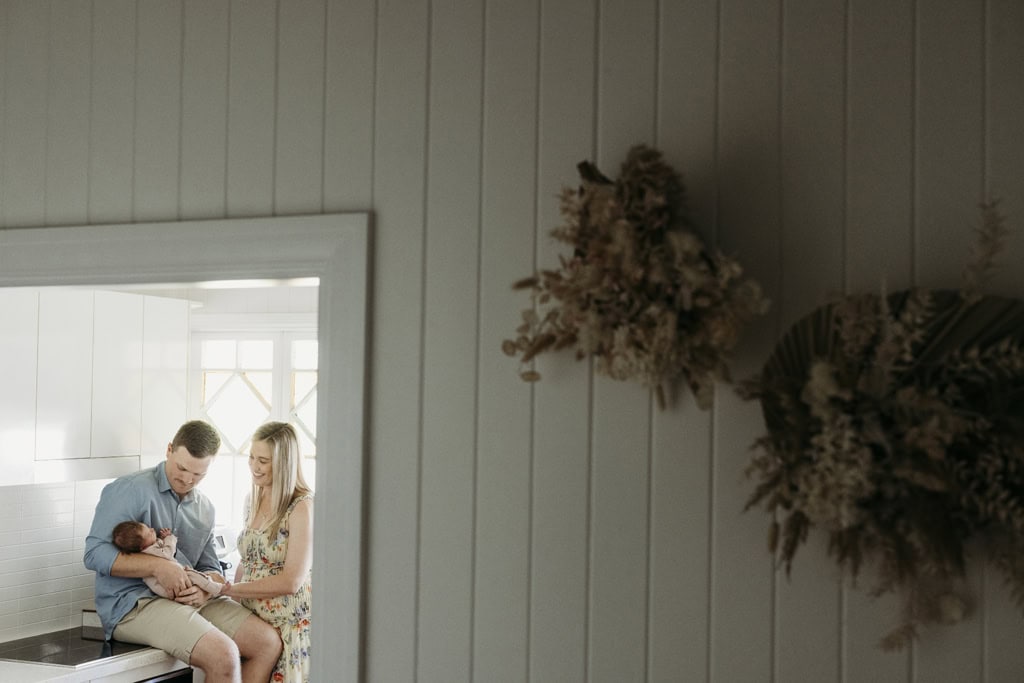 A couple gently cradles their newborn baby in their kitchen. They are framed by a doorway and in the foreground is dried flowers.