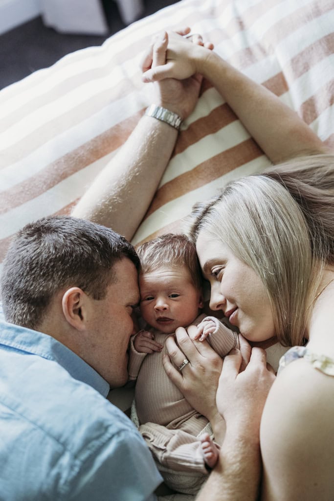 A couple hold hands as they lay down with their newborn between them. They are both smiling at her.