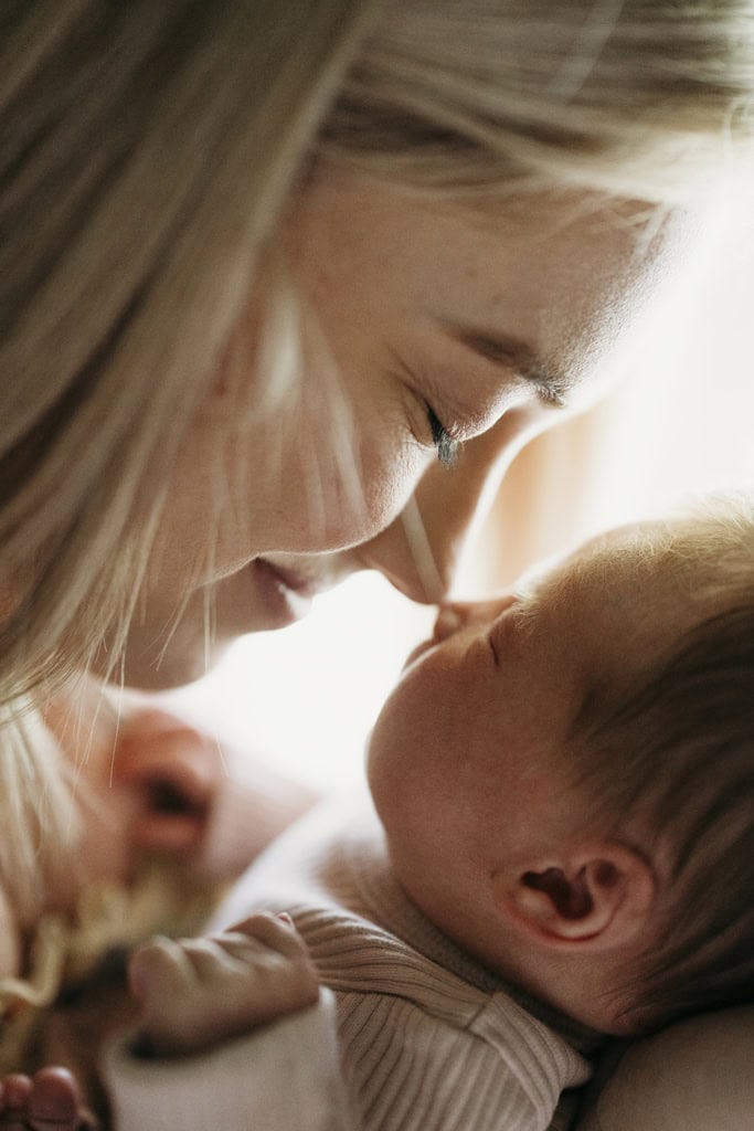 A mother gently touches her nose to her newborn's as she smiles. Light softly glows from behind them. 